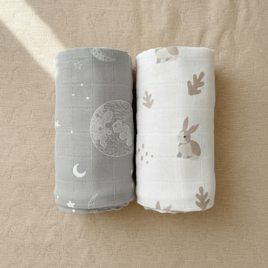 Two rolled-up bamboo swaddles bundle, one gray with celestial patterns and one white with bunny designs, on a beige surface.